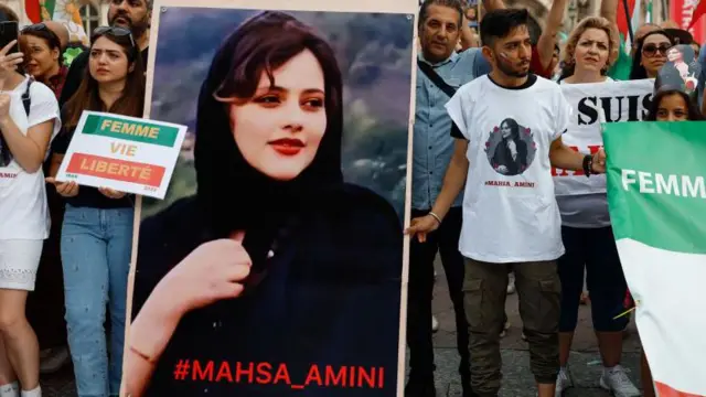 Una persona lleva una pancarta con la inscripción "Mahsa_Amini" en rojo y una imagen de Amini. La pancarta está rodeada de hombres y mujeres que visten camisetas con su imagen y portan banderas para conmemorar el primer aniversario de su muerte.