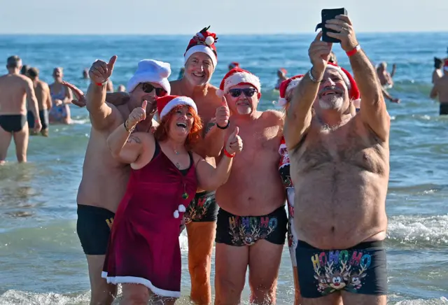 Un grupo de personas en el mar, con gorros de Papá Noel y trajes de baño, se toman una selfie con el pulgar hacia arriba.