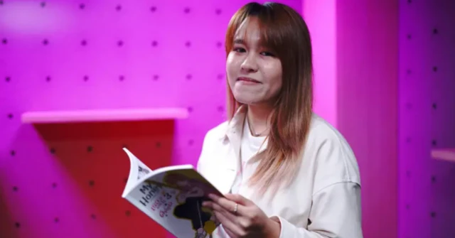 Ranuka Songmuang, vestida en camisa blanca, en un cuarto de paredes rosadas, sonríe a cámara mientras lee una revista manga