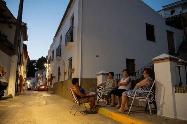 Unas señoras toman el fresco de la noche en una calle de Arriate, sentadas en unas sillas plegables mientras charlan.