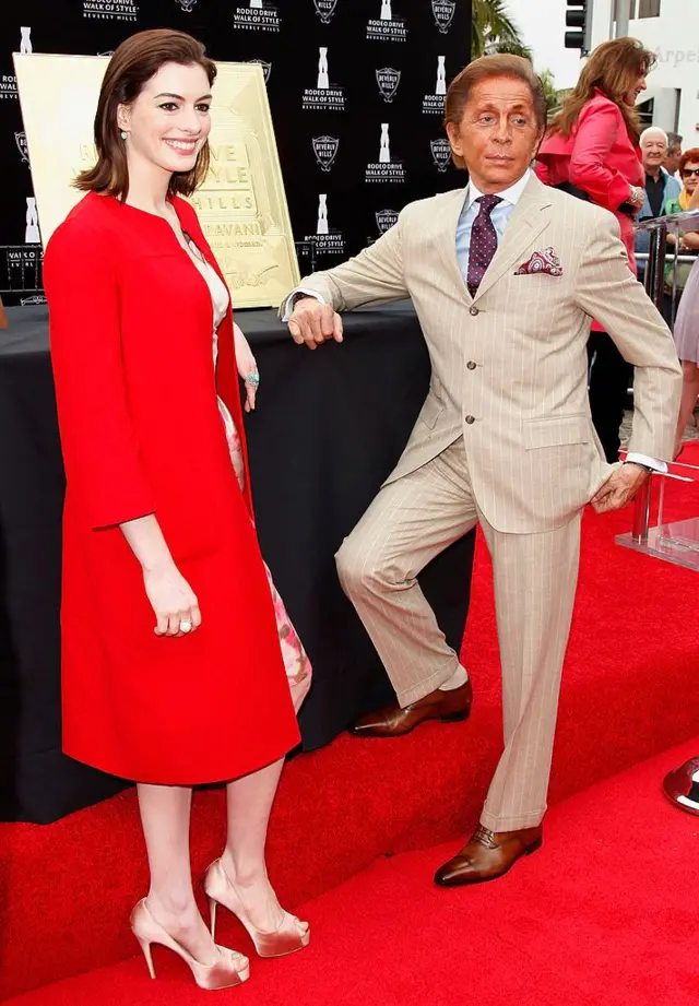 Anne Hathaway, vistiendo un abrigo rojo, posando para la prensa al lado de Valentino