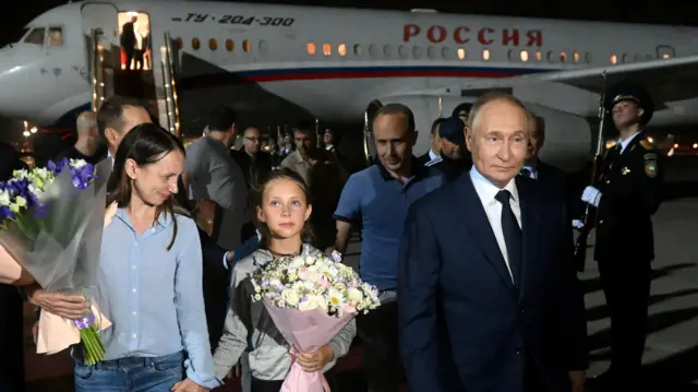 Una mujer de cabello largo sostiene la mano de una niña tras bajar de un avión. Ambas llevan ramos de flores. El presidente Putin está junto a ellas.