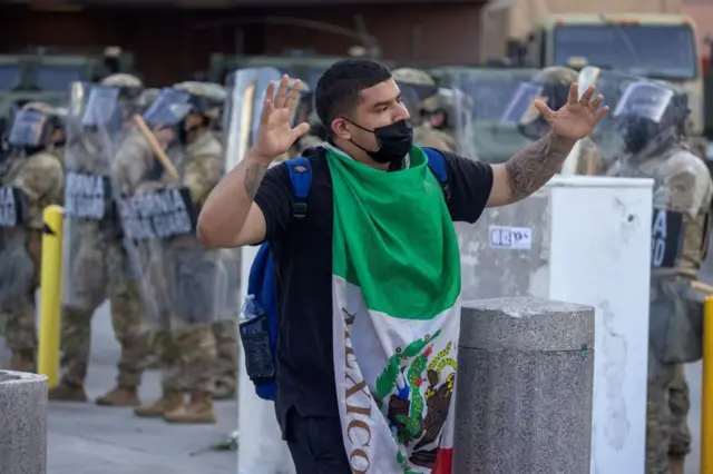 Un hombre envuelto en una bandera de México con los brazos en alto camina frente a soldados de la Guardia Nacional de California