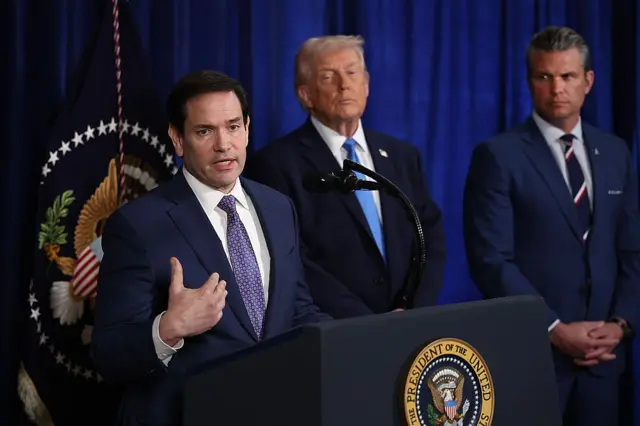 El secretario de Estado de EE. UU., Marco Rubio, habla durante una conferencia de prensa mientras el presidente estadounidense Donald Trump y el secretario de Guerra Pete Hegseth escuchan. Los tres van vestidos con traje y corbata. El fondo es una gran cortina de color azul y una bandera con el escudo de Estados Unidos.