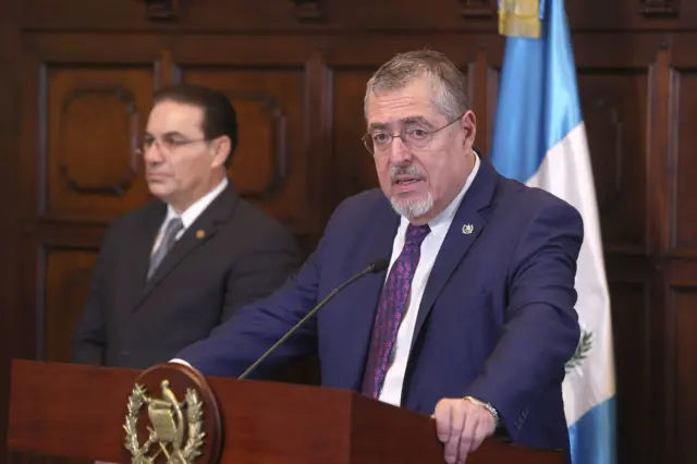 El presidente de Guatemala, Bernardo Arévalo, habla en un atril. Tiene la bandera de su país detrás y un hombre con traje oscuro en segundo plano a la derecha.