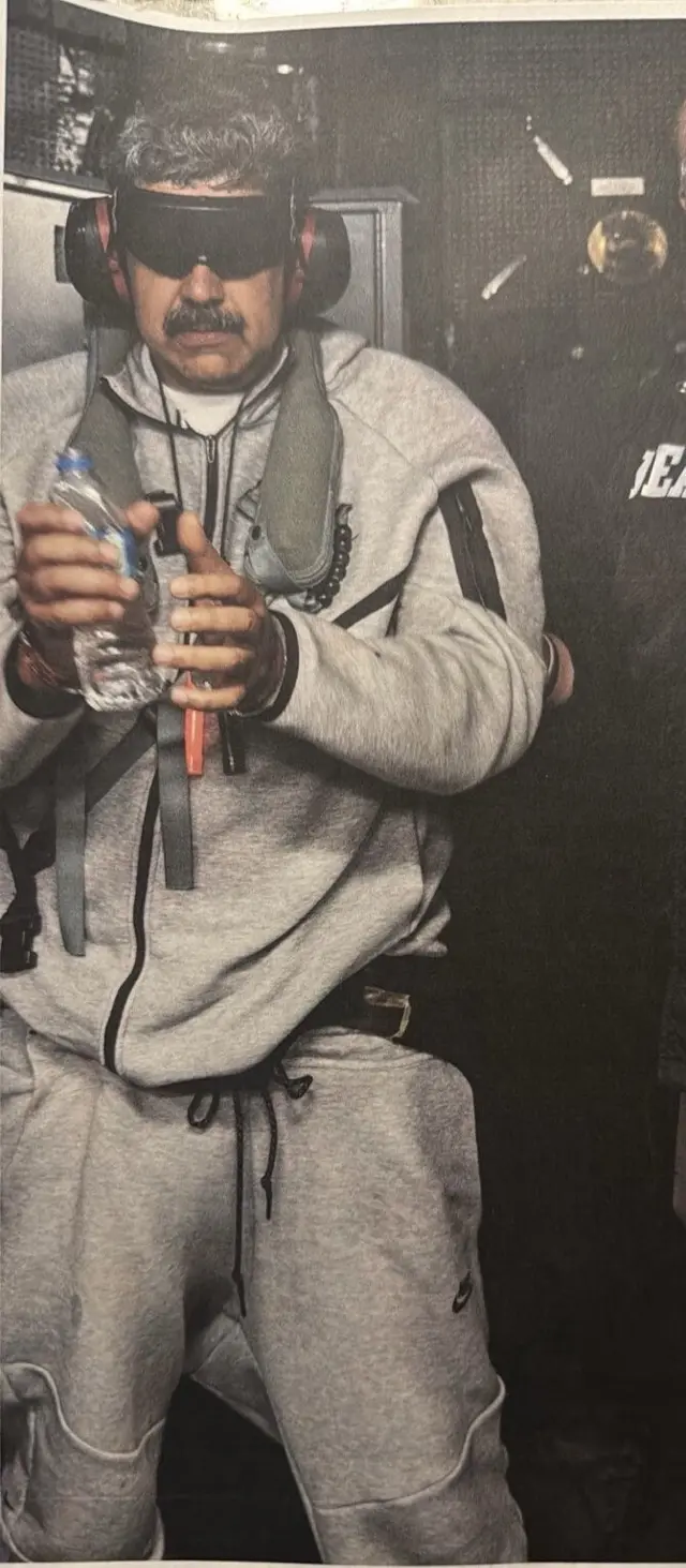 Cuerpo de Nicolás Maduro con anteojeras y protectores auditivos. Tiene las manos esposadas y sostiene con una de ellas una botella de plástico pequeña. Viste una camiseta blanca y un conjunto deportivo gris.