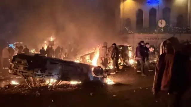 Autos en llamas y volcados en una calle por la noche. 