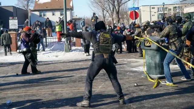 Un agente federal encapuchado, de espaldas a la cámara y con los brazos extendidos, está a punto de lanzar lo que parece ser una granada de gas lacrimógeno, mientras los manifestantes observan desde los porches cubiertos de nieve y otros agentes federales se encuentran detrás del agente.