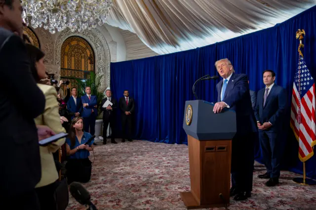 Donald Trump está parado frente a un atril con micrófono. Detrás está Marco Rubio y, junto a él, una bandera de Estados Unidos. De fondo se ven funcionarios y frente a Trump hay periodistas.
