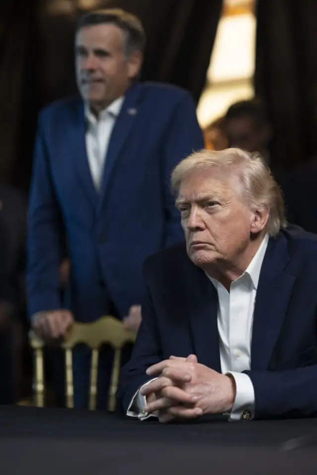 Ratcliffe parado detrás del respaldo de una silla y Trump sentado a la mesa.