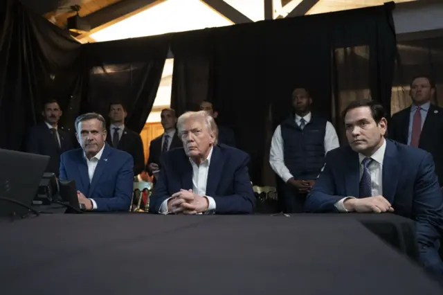 John Ratcliffe, Donald Trump y Marco Rubio sentados a la mesa, y asesores parados miran detrás.