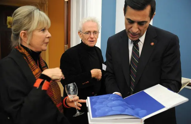 Daphne y Chuck visten de negro en la presentación de su libro, que se ve abierto en primer plano. Ella sostiene una copa de vino vacía mientras habla con el congresista Darrell Issa.