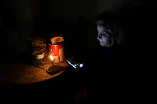 Una mujer sentada junto a una mesa con su rostro iluminado por una vela y el celular que sostiene en sus manos