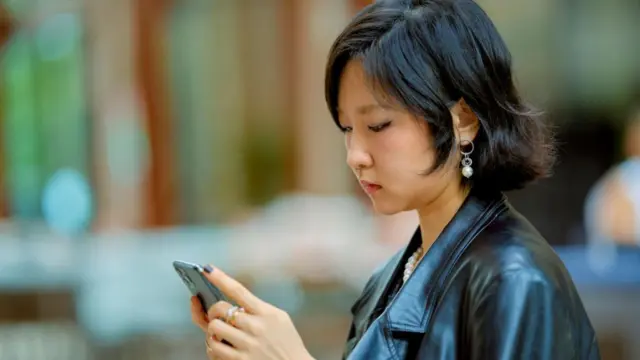 Una mujer joven mira su teléfono móvil mientras lo sostiene con ambas mano. Parece encontrarse en un espacio exterior.
