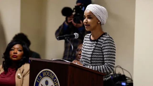 Una fotografía de Ilhan Omar dirigiéndose a una reunión municipal vestida con jersey de rayas negras y blancas, turbante blanco. Habla por un micrófono en un podio con otras personas en el fondo; el podio muestra un logotipo.