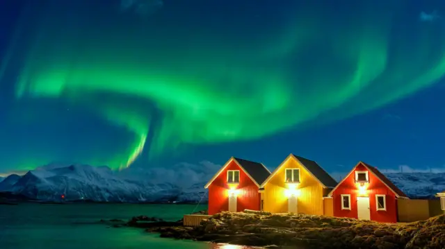 Aurora boreal sobre montañas nevadas y cabañas de pescadores junto al mar en Troms og Finnmark, Noruega