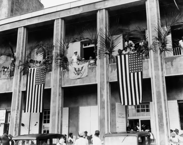 Foto en blanco y negro de la fachada de un edificio de St. Thomas con grandes banderas de Estados Unidos colgadas de sus balcones.