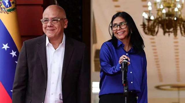 Jorge Rodríguez a la izquierda, vestido con chaqueta y camisa blanca, Delcy Rodriguez al lado, con camisa azul y pantalón negro, la mano en un micrófono, ambos sonríen. Al fondo se ve una estancia grande con una gran lámpara de techo y, a la izquierda, una bandera de Venezuela.