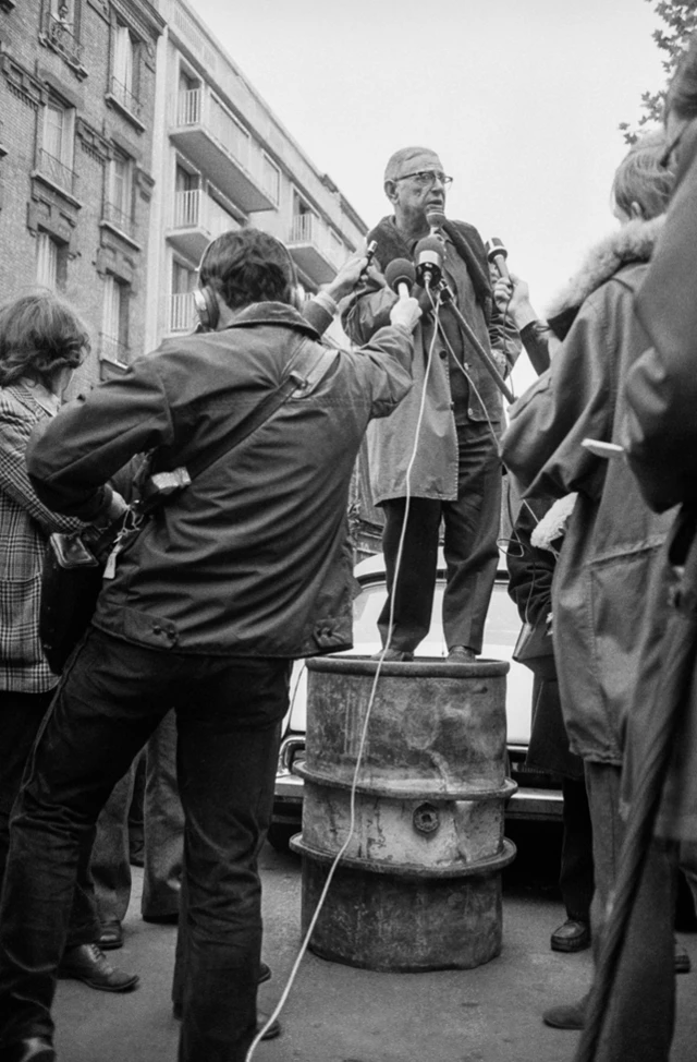 Sartre con un micrófono en la mano montado en un barril y rodeado de periodistas