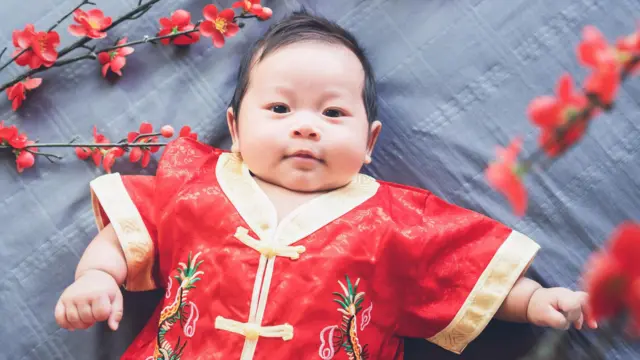 Un bebé vestido en un atuendo tradicional chino de color rojo con bordes dorados yace sobre un manto gris, rodeado de unas flores rojas