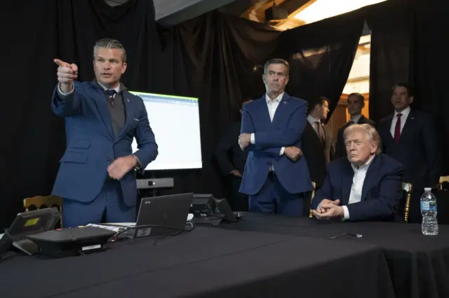 Hegseth está parado, apuntando hacia algo delante, mientras Ratcliffe y Trump miran lo que señala.