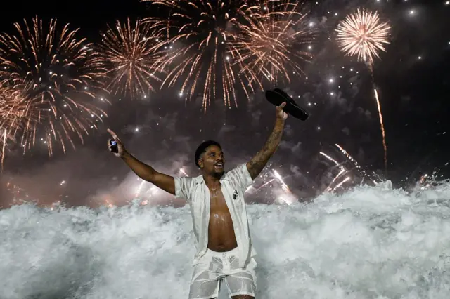 Un hombre celebra con los brazos en alto entre las olas del mar en Copacabana. En el fondo, el cielo está iluminado con fuegos artificiales