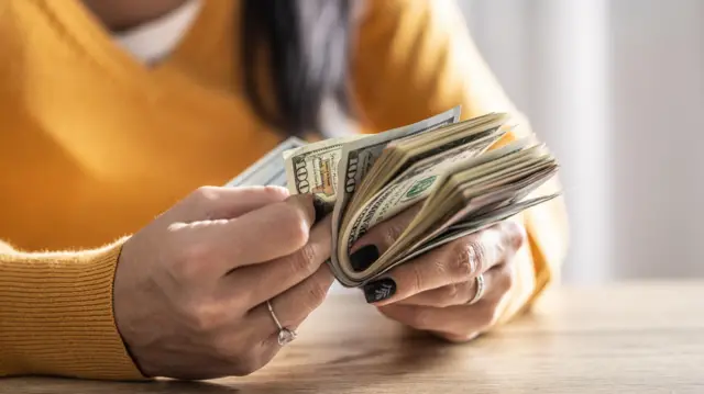 Primer plano de las manos de una mujer con un suéter amarillo, un anillo y las uñas pintadas de oscuro, contando muchos billetes de 100 dólares estadounidenses.