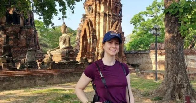 Luiza Z, una mujer brasileña, vistida con una playera color vino tinto, con una gorra de béisbol y una cámara al hombro, sonríe frente a las ruinas del templo de un Buda en Tailandia