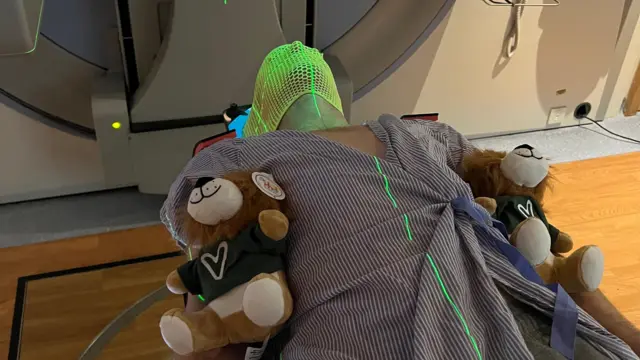 Anthony Perriam está acostado en una cama de hospital con dos ositos de peluche a su lado. Lleva puesta una mascarilla porque le van a realizar una exploración para detectar cáncer.