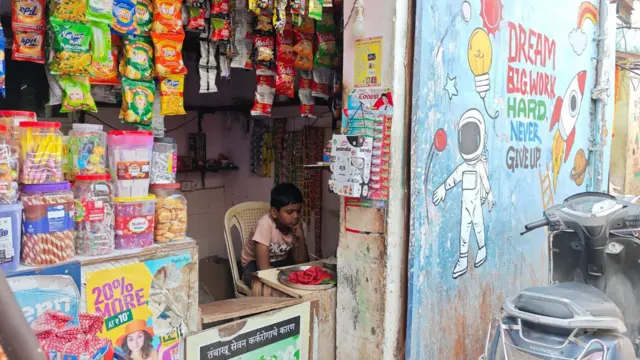 Un niño está sentado en una tienda mirando su teléfono. Afuera, un mural dice: "Sueña en grande, trabaja duro, nunca te rindas".
