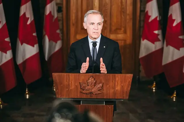 Mark Carney dando un discurso en un podio, rodeado de banderas de Canadá.
