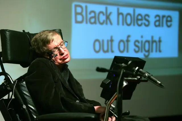 Stephen Hawking, vestido de negro, en su silla de ruedas, frente a un monitos pequeño y en el fondo una pantalla gigante dice: Black holes are out of sight