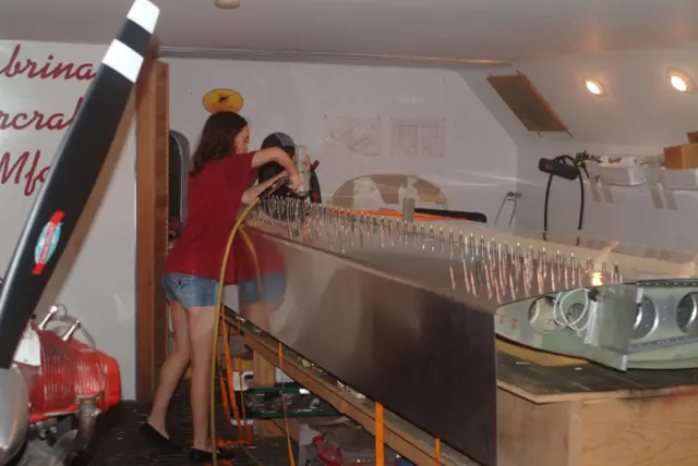 Sabrina González Pasterski, con una camisa roja y unos shorts de jeans, trabajando unas partes metálicas con una herramienta conectada a un cable