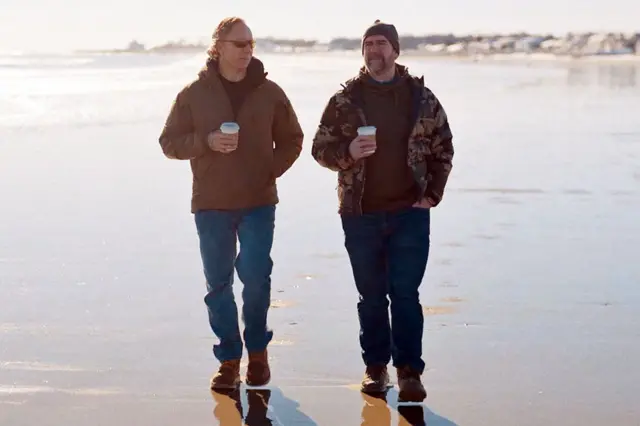Greg Squire y Pete Manning caminando por la playa con bebidas para llevar. Ambos llevan pantalones azules y chaquetas marrones.