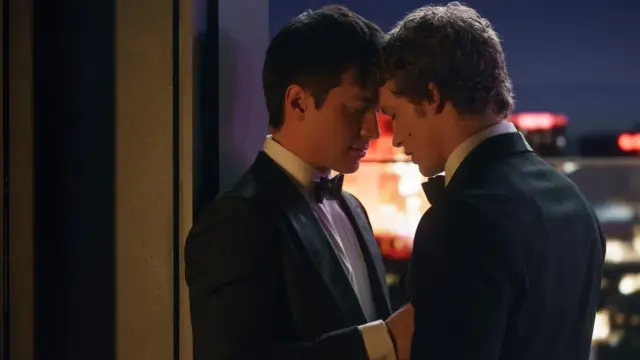 Shane Hollander, interpretado por Hudson Williams, e Ilya Rozanov, interpretado por Connor Storrie, ambos vestidos con esmoquin, se abrazan en una azotea en una escena de Heated Rivalry ("Más que rivales").