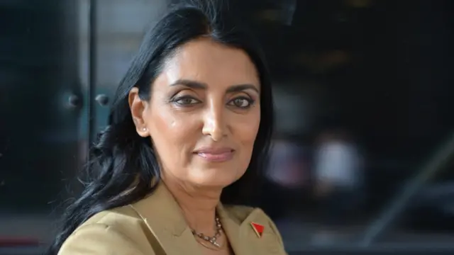 Aneeta Prem mira directamente a la cámara. Tiene el pelo oscuro y viste una chaqueta de traje de color claro. Lleva una insignia roja y negra en la solapa, en forma de triángulo.