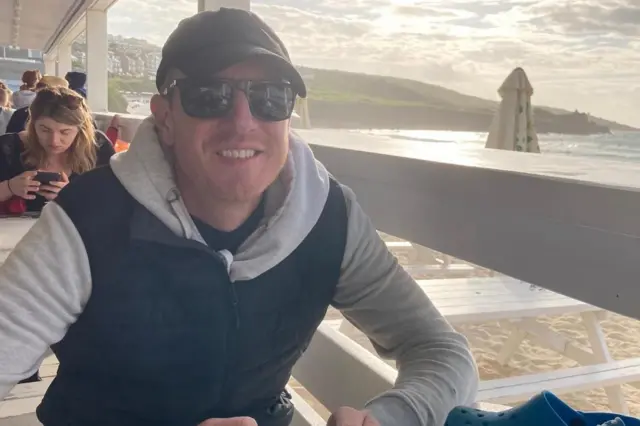 Patrick sonriendo y sentado en un bar frente a la playa, con gafas de sol.