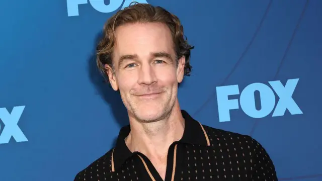 James Van Der Beek en una alfombra roja. Sonríe con un fondo azul detrás.