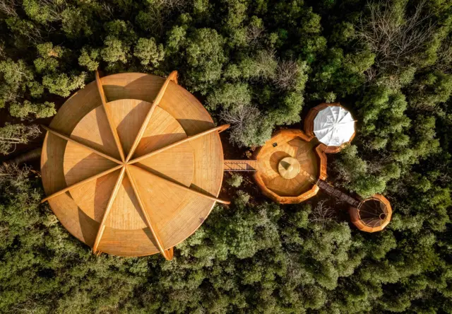 Vista cenital de in gran edificio circular en madera, que está conectado a otros dos espacios circulares -también en madera- a través de puentes que conectan toda la estructura.