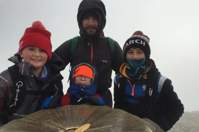 Gerwyn y sus tres hijos llevan gorros de lana, abrigos y mochilas. Están de pie junto al montículo en la cima de Yr Wyddfa. Los niños son adolescentes o incluso menores. Todos sonríen. La visibilidad es nula tras ellos debido a un muro de nubes grises.