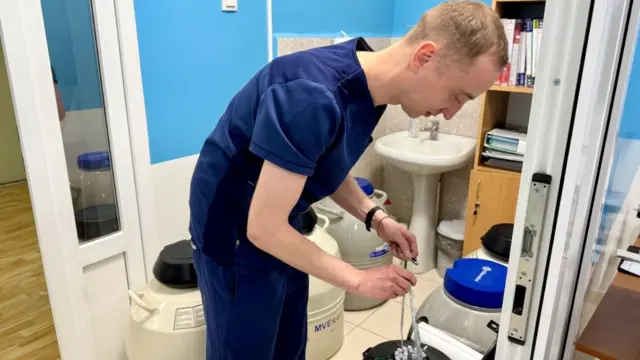 Un hombre con uniforme azul usa una pipeta en el Centro de Medicina Reproductiva estatal de Kiyv. Detrás de él hay enormes cubas blancas.