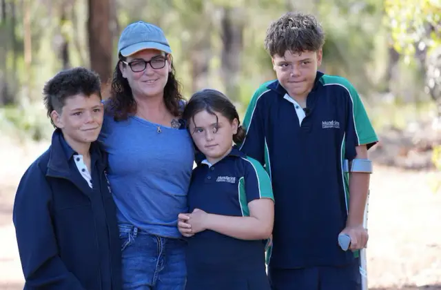 Beau está de pie junto a su madre, con una camiseta azul, y su hermana pequeña Grace. Todos los niños llevan uniforme escolar. Austin está de pie con muletas.
