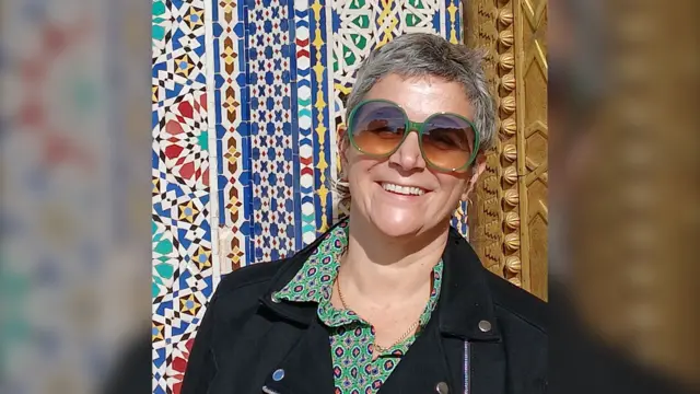 Raquel, con unos lentes de sol de montura verde, sonríe frente a la cámara. Detrás de ella hay un mosaico colorido. Su cabello es corto y viste una blusa verde y una chaqueta negra