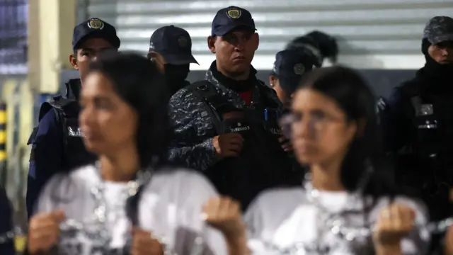 Unas madres de detenidos cerca de un puesto policial en Caracas y al fondo policías antimotines los vigilan.