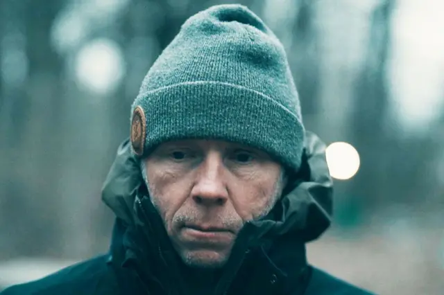 Retrato de Greg Squire paseando por el bosque, en el que lleva un abrigo oscuro y un gorro gris.