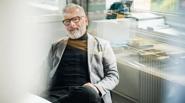 Un hombre, posiblemente un baby boomer, sentado en su oficina con gafas de marco negro, un cuello tortuga negro, una chaqueta gris. Tiene las manos sobre las piernas.