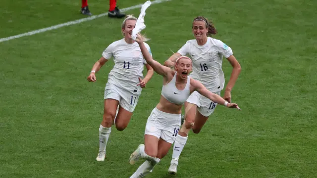 Chloe Kelly se quita la camisa mientras sonríe y corre por la cancha de fútbol, seguida de dos compañeras.