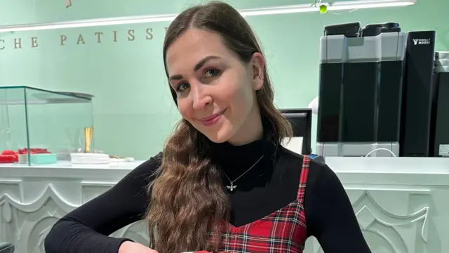 Una mujer con el pelo largo y castaño, vestida con un jersey negro de cuello alto y un vestido a cuadros, sonriendo.