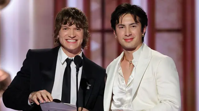 Connor Storrie, sonriendo y sosteniendo un sobre, vestido con un traje oscuro y corbata, y Hudson Williams con una camisa blanca de cuello abierto y un traje blanco, en el escenario de los Globos de Oro.