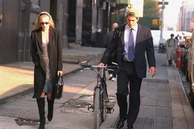 Carolyn Bessette y John F. Kennedy caminan por una calle. Ella lleva unos lentes de sol, viste unas botas largas negras, un vestido corto y un sobretodo negro. El tiene una gorra de beisbol volteada hacia atrás, viste un traje negro, con camisa azul y corbata y arrastra con su mano derecha una bicicleta.